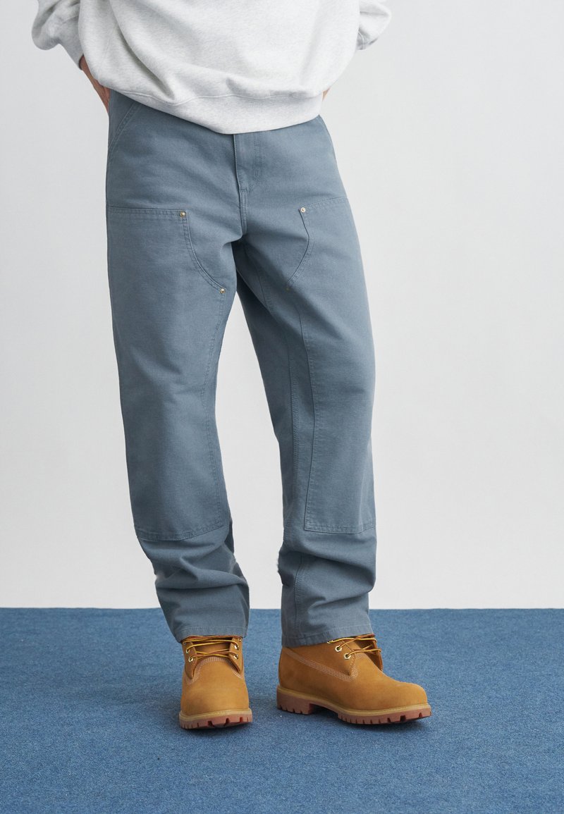 Άτομο που φοράει ανοιχτό μπλε εργασιακό παντελόνι με μπροστινές τσέπες και μπεζ μπότες με κορδόνια, στέκεται πάνω σε μπλε χαλί με τα χέρια πίσω από την πλάτη.