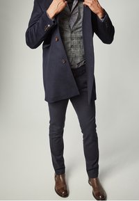 Man som justerar en mörk marinblå kappa över en grå rutig väst, iförd mörka byxor och bruna läderstövlar, stående mot en ljusgrå bakgrund.