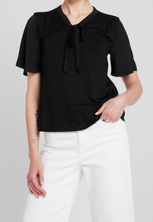 Frau trägt eine schwarze kurzärmelige Bluse mit Bindedetail vorne und weiße hoch taillierte weite Hose, Hand in der Tasche.