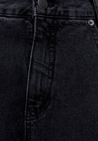 Tejido de mezclilla negra con una textura sutil, que presenta un cierre frontal con cremallera, un botón y un contorno de bolsillo curvado en el lado derecho.