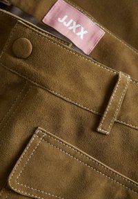 Tissu en daim brun avec des coutures visibles et une poche carrée. Présente une étiquette rose avec du texte blanc. Fermeture par bouton à la taille.