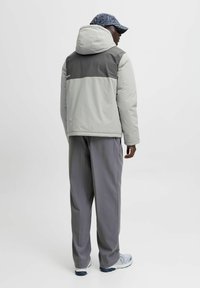 Helle graue gepolsterte Jacke mit einem dunklen grauen Schulterbereich, Kapuze und elastischen Bündchen, kombiniert mit lockeren grauen Hosen und einer gemusterten Mütze.