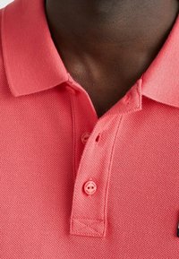 Rosa pikétröja med strukturerad tyg, två knappar vid kragen och en liten svart accentetikett nära fållen.