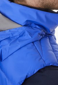 Blå pufferväst med grå detaljer, som visar en kviltad textur och hög krage. Har en slät, glänsande ytfinish och vadderad design.