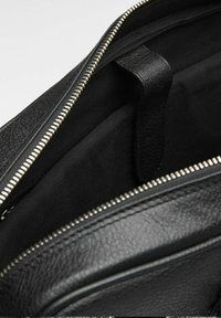 Indersiden af en sort lædertaske med en struktureret finish, der har en lynlås lukning og en stofforet. Indeholder en lille stofetiket.