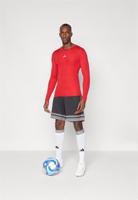 Rød langærmet atletisk top, grå shorts med sort og hvid striber, hvide sokker og støvler; holder en multicolor fodbold.