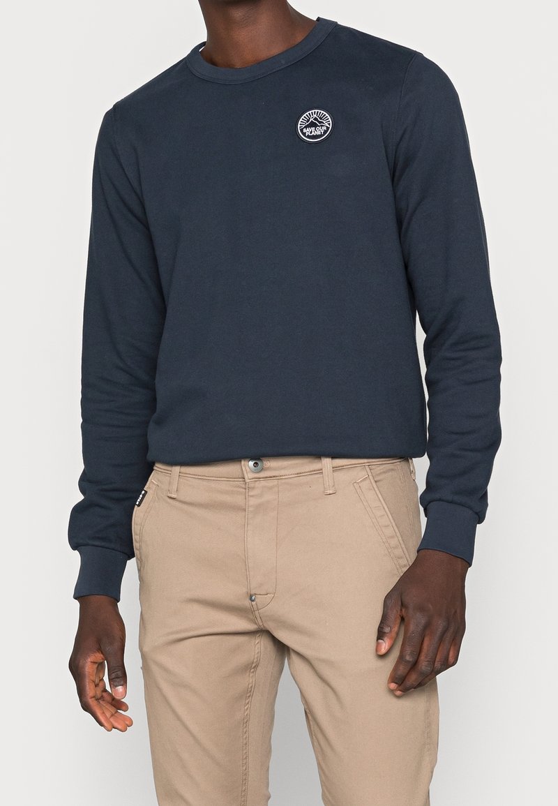 Homme portant une chemise bleue foncée à manches longues avec un écusson « Save Our Planet » et un pantalon beige, debout devant un fond uni.