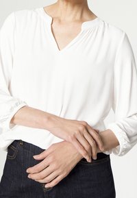 Blusa bianca con scollo a V e dettaglio arricciato al collo, caratterizzata da maniche lunghe con polsini elastici. Indossata con jeans scuri.