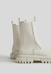 White ankle boots with elastic side panels, rubber soles, and pull tabs. Smooth exterior with textured accents, featuring a chunky tread.