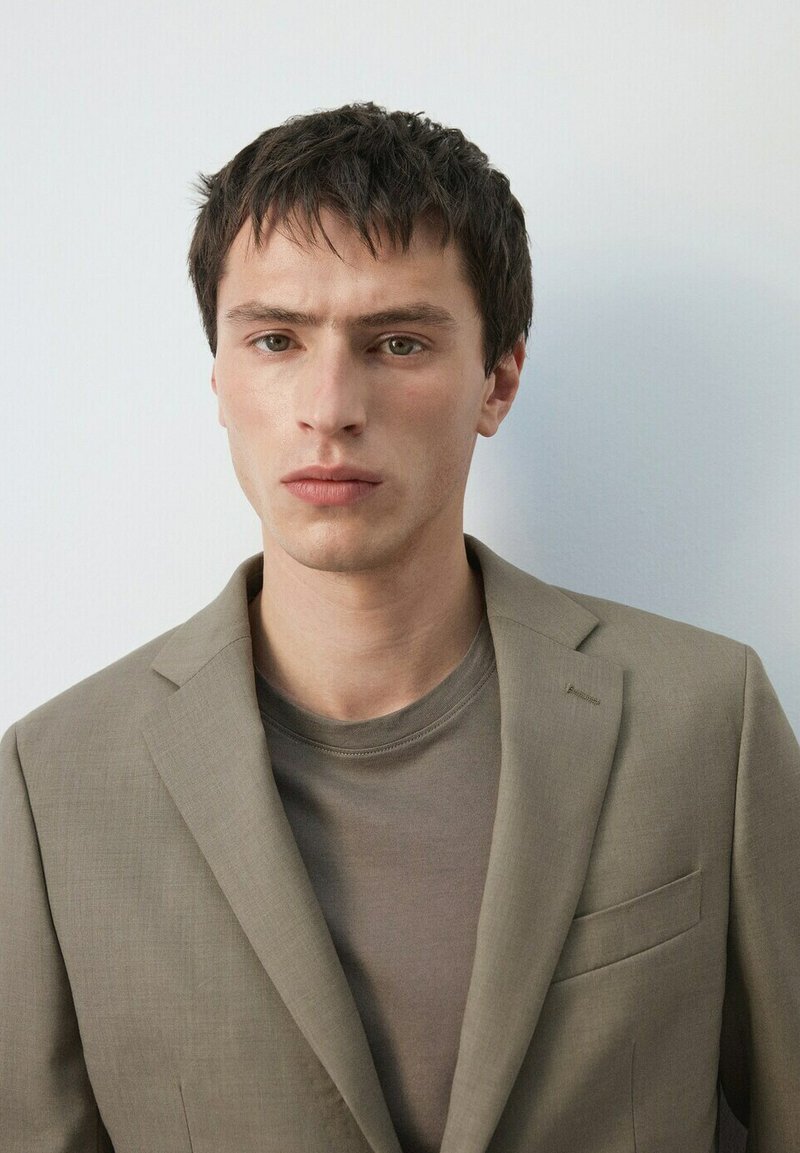 Jonge man met kort donker haar, gekleed in een beige blazer over een taupe ronde hals shirt, met een neutrale uitdrukking, staande tegen een lichtgrijze achtergrond.