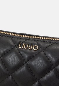Bolso de piel acolchada negra con cremallera dorada y detalles de logo. Presenta un acabado texturizado y un diseño estructurado.