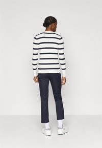 Fehér és tengerészkék csíkos bordás pulóver hosszú ujjal, tengerészkék nadrággal és fehér sportcipővel párosítva. A hátsó nézet kiemeli a ruhadarab részleteit.