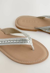 Zilveren flip-flops met metalen kralenaccenten op een gestructureerde voetbed, met een platte, tan kleurige zool en een T-band ontwerp.