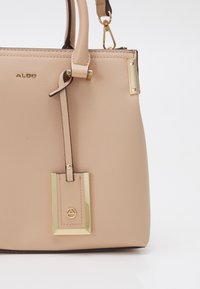 Sac à main synthétique beige avec une finition texturée, mettant en avant des accents en métal doré et une étiquette amovible. Bandoulière réglable incluse.