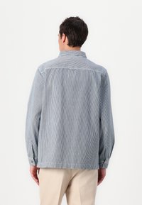Casaco de ganga riscado em azul claro e branco, com colarinho, punhos com botões e um corte descontraído. Costuras visíveis como destaque.
