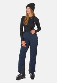 Junge Frau trägt eine schwarze Mütze, ein schwarzes Langarmoberteil, marineblaue Schneehose mit Trägern und schwarze Ski-Stiefel und steht vor einem weißen Hintergrund.