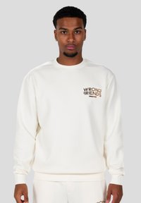 Sudadera blanca de canalé con un pequeño logo marrón "WRONG FRIENDS" en el pecho. Cuello redondo y mangas largas.