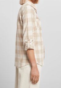 Donna che indossa una camicia a maniche lunghe beige e bianca a scacchi con le maniche arrotolate e pantaloni bianchi, in piedi davanti a uno sfondo neutro.