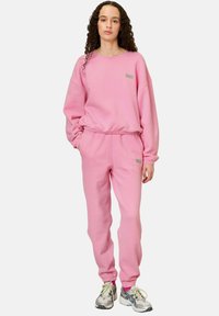 Sweatshirt en polaire rose et pantalon de survêtement assorti avec une coupe décontractée. Présente un détail de logo vert sur la poitrine et à la cheville. Tissu texturé.