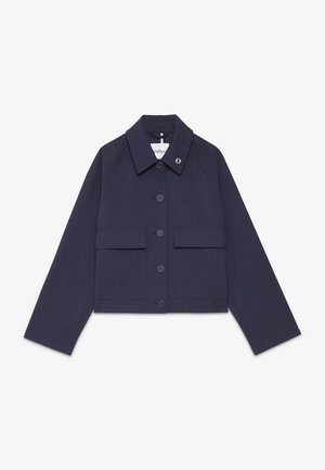 Donna con capelli scuri raccolti in uno chignon basso che indossa una giacca blu navy con bottoni e pantaloni neri ampi con piccoli motivi rossi su uno sfondo semplice.
