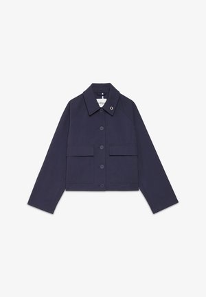 Donna con capelli scuri raccolti in uno chignon basso che indossa una giacca blu navy con bottoni e pantaloni neri ampi con piccoli motivi rossi su uno sfondo semplice.