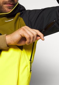 Giacca multifunzionale da uomo con parte superiore nera e sezione inferiore gialla, tasche con zip e polsini a coste. Tessuto testurizzato.
