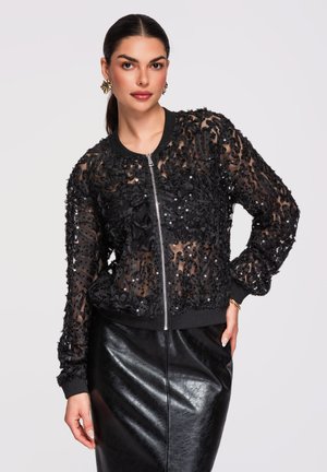 Femme aux cheveux foncés portant une veste bomber noire transparente avec des sequins floraux et une jupe crayon en cuir noir, posant devant un fond uni.