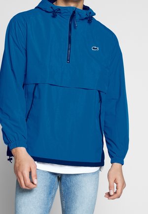 Uomo che indossa una giacca con cappuccio blu con mezza zip, polsini elastici e logo Lacoste, abbinata a jeans azzurri e una camicia bianca sotto.
