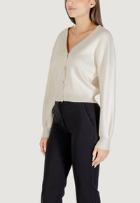 Cárdigan de canalé crema con escote en V, mangas largas, cierre de botones y un diseño corto. Combinado con pantalones negros para crear contraste.