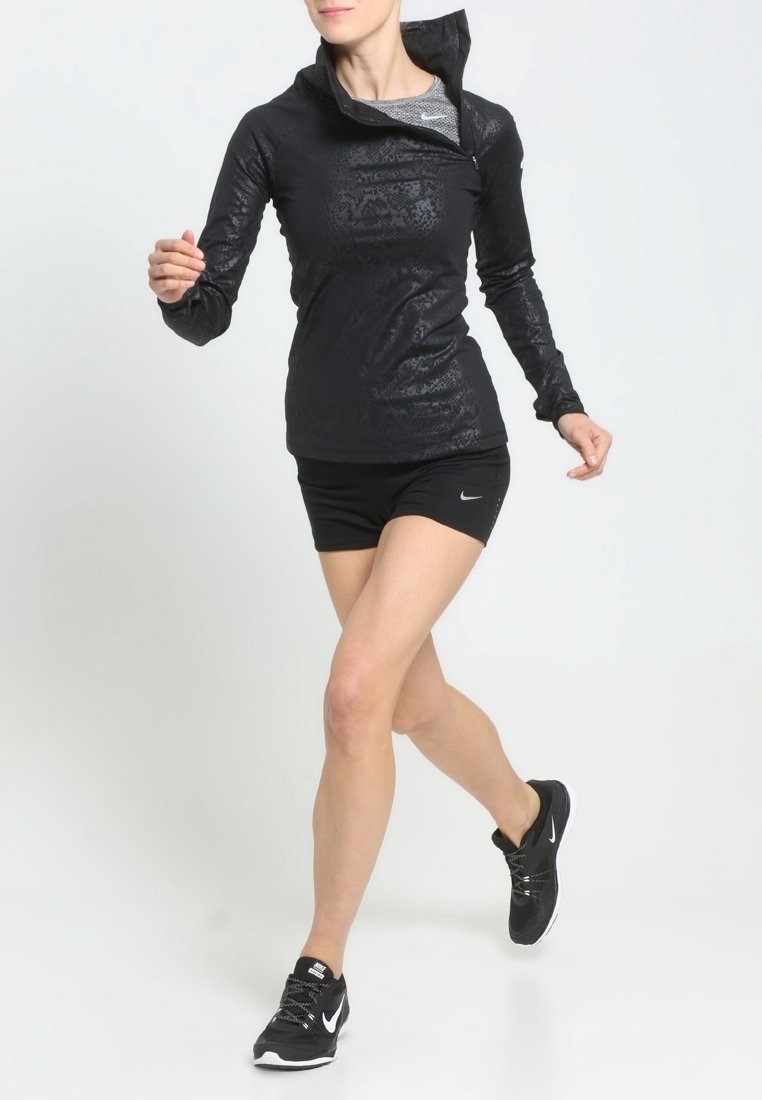 Mujer con capucha atlética negra, pantalones cortos y zapatillas de correr caminando o trottando frente a un fondo blanco liso.