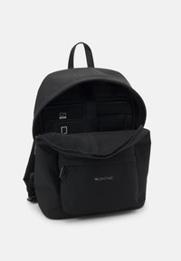 La mochila negra de nailon cuenta con un bolsillo frontal con cremallera, correas ajustables y un logotipo en relieve. El interior incluye secciones acolchadas para la organización.