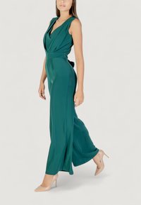 Modelo con un mono verde de escote en V profundo y piernas fluidas, caminando elegantemente mientras lleva tacones nude sobre un fondo claro.