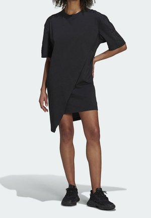 Mujer con vestido tipo camiseta negro asimétrico de manga corta y zapatillas negras, de pie con una mano en la cadera sobre un fondo liso.