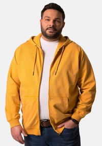 Sudadera amarilla con cremallera hecha de tejido suave, con dos bolsillos frontales, cordones ajustables y puños acanalados. Llevada sobre una camiseta blanca.