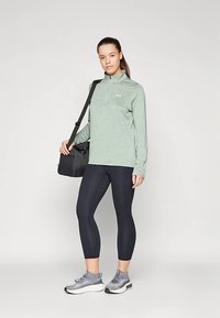 Lichtgroen sportief shirt met lange mouwen, zwarte legging, grijze sneakers. Model draagt een zwarte tas. Soepele stof met een aansluitend ontwerp.