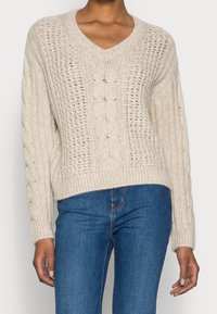 Pull beige en maille avec col en V, coupe ample, motifs torsadés détaillés et poignets côtelés, associé à un jean ajusté bleu foncé.