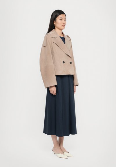 Veste cropped en suède beige à double boutonnage avec de grands boutons noirs, associée à une jupe longue navy et des mules blanches. Texture lisse.