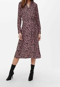 Abito rosa a maniche lunghe con strisce zebrate nere, chiusura a bottoni sul davanti, vita accentuata da una cintura e lunghezza midi, abbinato a stivali neri con tacco.