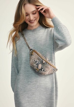Torebka crossbody w imitacji węża w beżowych i brązowych odcieniach z złotym zamkiem. Krzywizna i regulowany pasek. Noszona na szarym swetrze.