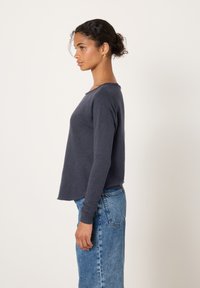 Moteris su garbanotais plaukais, surištais atgal, dėvinti tamsų ilgomis rankovėmis marškinėlį ir mėlynas džinsus, stovinti profilio pozoje priešais paprastą šviesų foną.