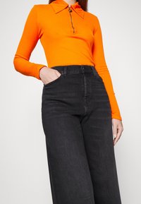 Femme portant un haut à manches longues orange vif avec une fermeture éclair à l'avant et un jean noir taille haute à jambes larges, une main dans la poche.