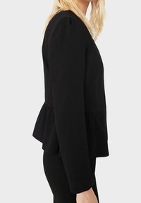 Blonde vrouw in een aansluitend zwart langemouwtop met een peplumrand, staand in profiel tegen een lichte achtergrond.