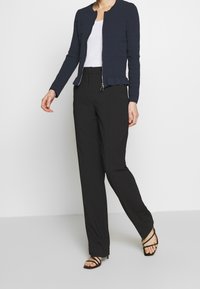 Námořnický texturovaný kabát s kulatým výstřihem a zipem, v kombinaci s černými kalhotami s širokými nohavicemi a černými páskovými podpatky.