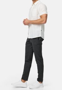 Mężczyzna w białej, krótkosleewej koszuli zapinanej na guziki, ciemnoszarych spodniach i białych sneakersach, stoi z rękami złożonymi przed sobą.