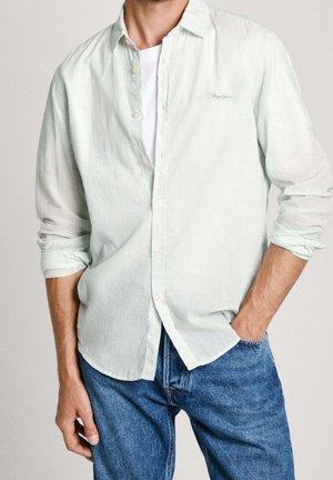 Homme portant une chemise boutonnée vert clair sur un t-shirt blanc aux manches retroussées et un jean bleu, une main dans la poche.