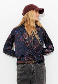 Blouse florale sombre en marine, ornée de motifs violets et verts, assortie à un jeans en denim gris taille haute et une casquette en velours côtelé bordeaux.