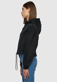Felpa nera corta con cappuccio regolabile, ampio cappuccio e maniche oversize, abbinata a jeans blu a vita media. Tessuto morbido e vestibilità rilassata.