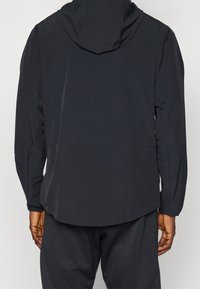 Homme portant une veste à capuche noire et un pantalon noir assorti, vu de dos sur un fond uni.