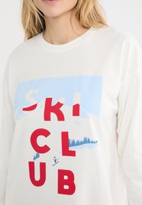 Biała koszulka z długim rękawem z niebiesko-czerwonym nadrukiem graficznym, który zawiera stylizowany napis „SKI CLUB” oraz narciarza na stoku z górami w tle.