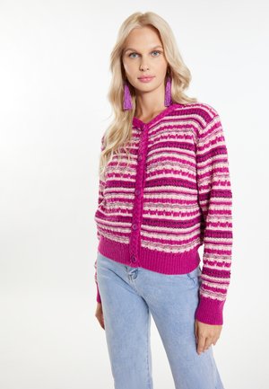 Femeie blondă purtând un cardigan cu dungi roz și albe cu nasturi și blugi albastru deschis, stând pe un fundal alb simplu.